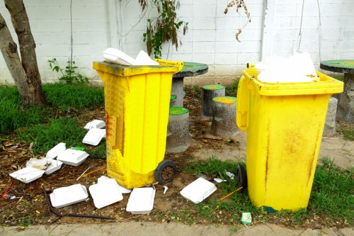 Two yellow wheeled rubbish bins made of plastic with textured surfaces are positioned outdoors on a grass verge beside a concrete pavement, against a white painted wall. One bin has its lid partially open, revealing white disposable food containers and paper packaging inside, while the other is closed with some waste visible around its base. Several discarded white foam takeaway containers and small bits of litter are scattered on the ground nearby, some leaning against the bins and others lying on the grass and pavement. Behind the bins, there are small round stools with green and yellow tops arranged in a slight arc, and a black round table is partially visible. The scene suggests an informal area where refuse has been left uncollected, illustrating a typical roadside rubbish issue in a residential or commercial neighbourhood, possibly in London, with some greenery and a tree trunk supporting the setting. The environment highlights the importance of waste management services like those offered by Commercial Waste Notting Hill.