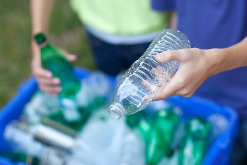 A close-up of a person's hand holding a clear plastic water bottle with a ridged screw cap, positioned in the foreground of the image. In the background, two individuals are visible, one in a light green top and the other in a blue top, standing outdoors on a grassy area. They are near a blue recycling bin filled with various disposed plastic bottles and waste materials, indicating a rubbish collection or recycling activity. The scene suggests an environmental cleanup in a residential or public space, consistent with waste management services offered in Notting Hill. The plastic bottles are transparent and green, with some caps visible, and the environment is well-lit with natural daylight, emphasizing the focus on waste sorting and disposal.