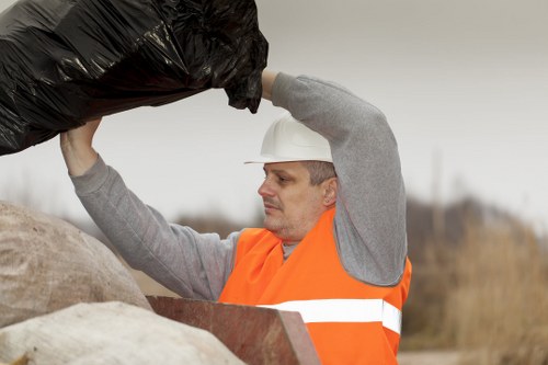 A male waste collection worker wearing a white safety helmet and an orange high-visibility vest over a grey long-sleeved top is seen lifting a large black plastic rubbish bag above his head. He is in an outdoor environment with a cloudy sky and what appears to be a rural or suburban area in the background. The worker is standing next to a pile of similar rubbish bags and debris, which include beige and brown materials of different textures, possibly fabric or construction waste, placed on the ground or a flat surface. The scene emphasizes the manual handling of waste, relevant to commercial waste removal services in the Notting Hill area, and showcases the worker preparing for rubbish collection or disposal. The environment suggests a typical waste management operation in an open space, with natural light highlighting the textures of the materials and the safety gear worn by the worker, aligning with the services provided by Commercial Waste Notting Hill.