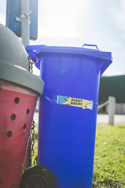 A close-up view of a blue plastic rubbish bin with a lid slightly open, positioned outdoors on a grassy area. The bin features a small, torn yellow and blue sticker with the partially visible message