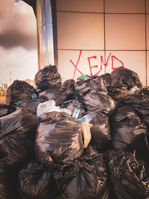 A large pile of black rubbish bags, many of which are tied at the top and appear full, are stacked against a modern building with a tiled exterior. Several light blue plastic water bottles are visibly